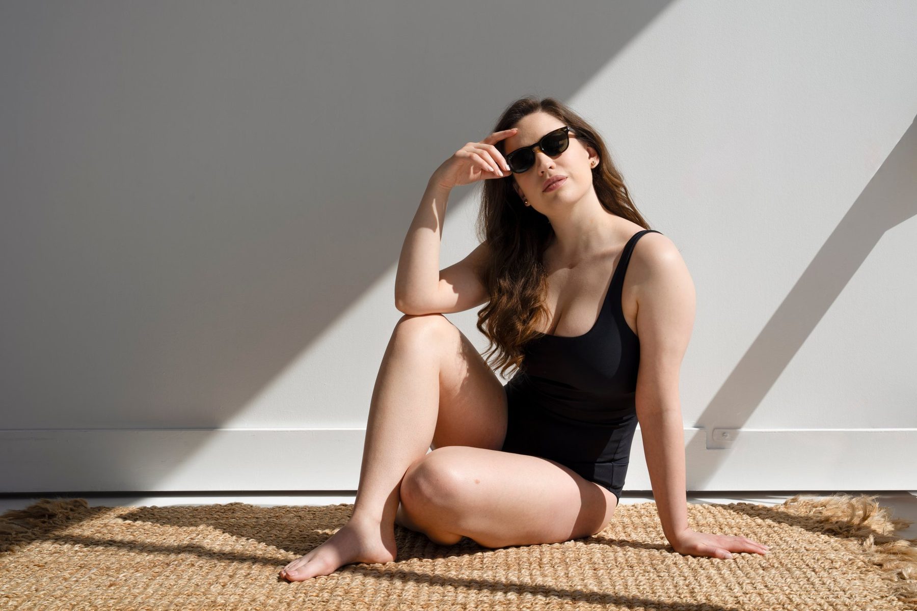 Model in a black one-piece and sunglasses seated on a jute rug