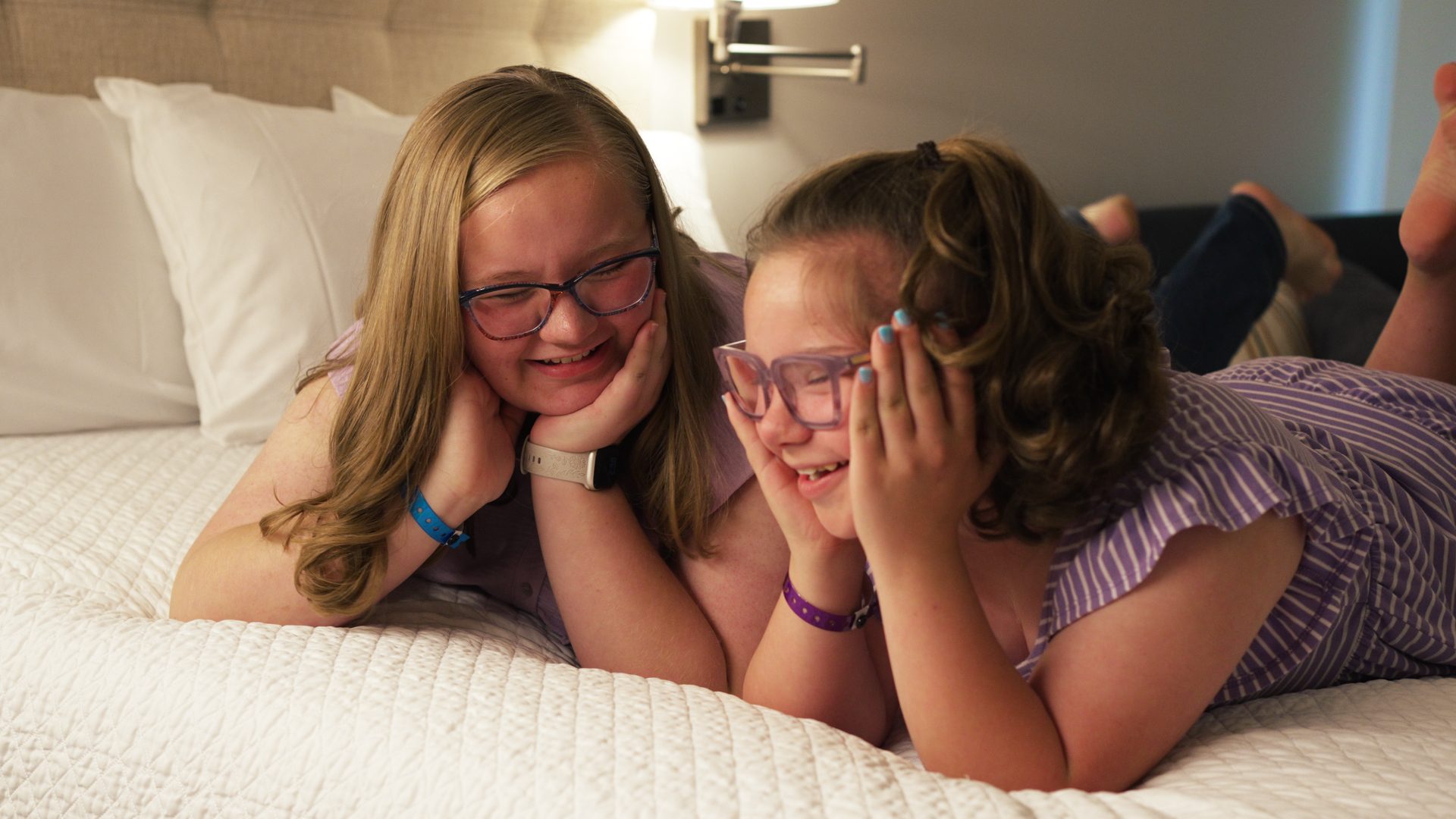 Two sisters smiling on a bed in a family room