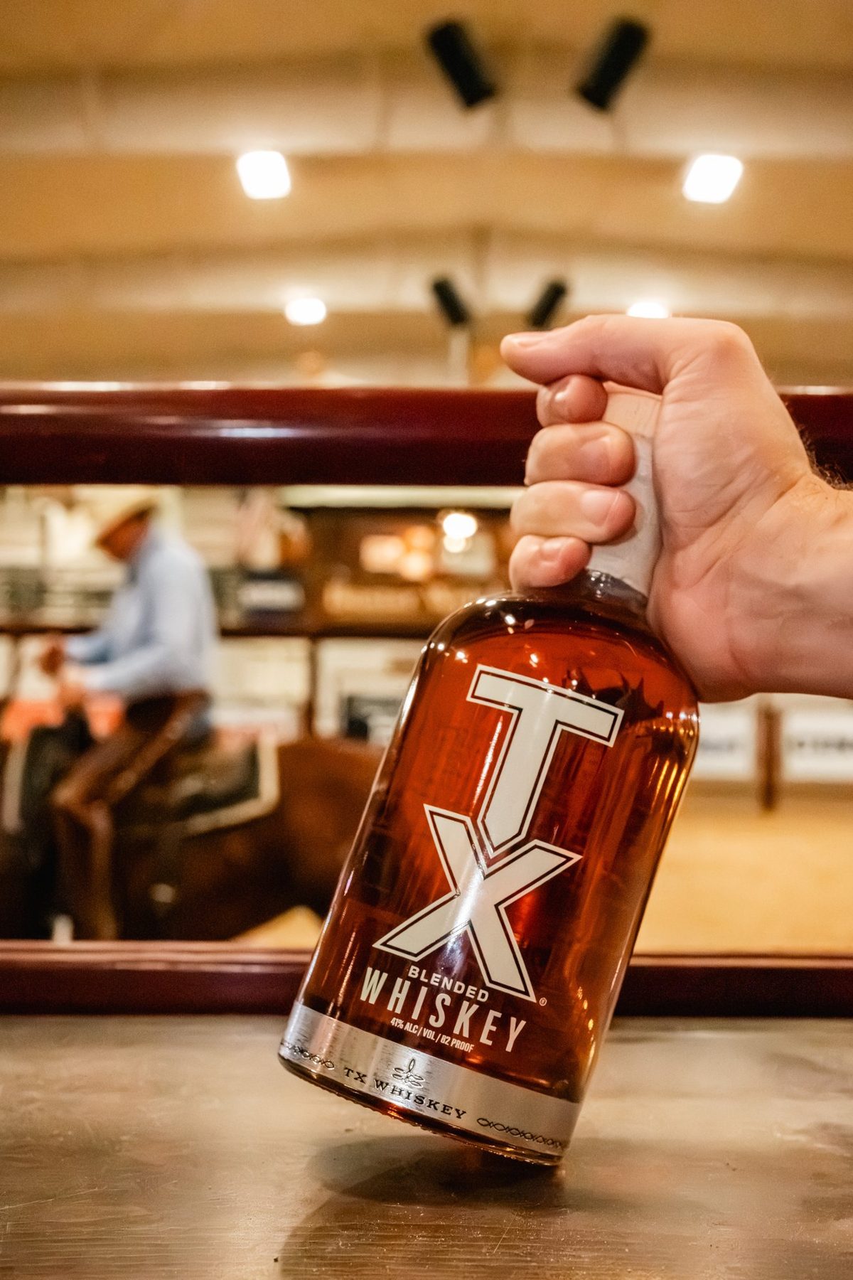 Hand holding a TX Whiskey bottle over a rodeo arena
